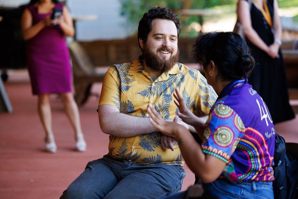 Two people in patterned shirts engage in a conversation while sitting outdoors, with others standing in the background.