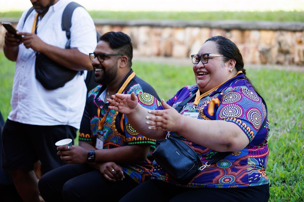 Two people in patterned shirts sit outdoors, laughing and clapping, with a third person standing nearby on the left.