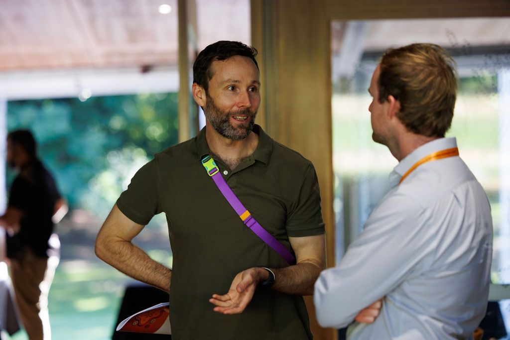 Two people in casual attire converse in an indoor space with large glass windows, one gesturing while speaking.