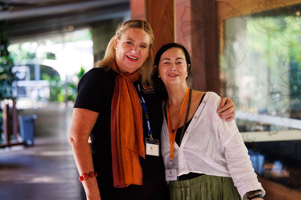 Two people with warm smiles stand side-by-side in a corridor, wearing colorful scarves and lanyards over casual clothing.