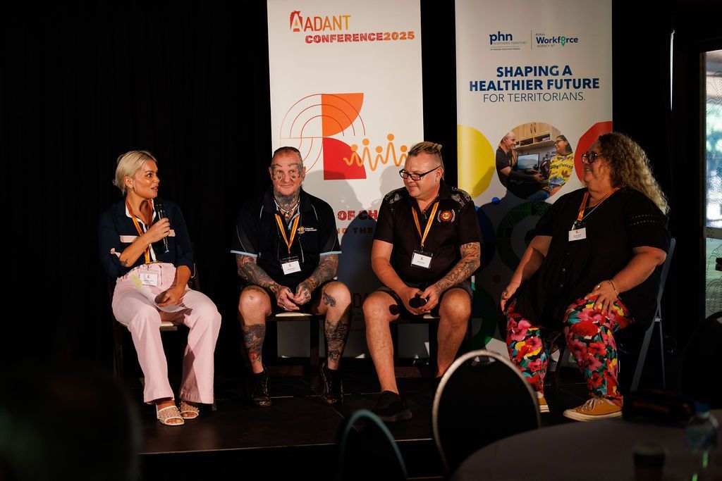 Four people seated on chairs during a panel discussion on a stage with banners, engaged in conversation.