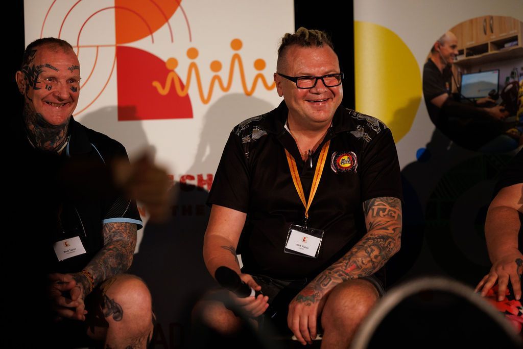 Two individuals with tattoos sit on a stage during an event, smiling while participating in a panel discussion.