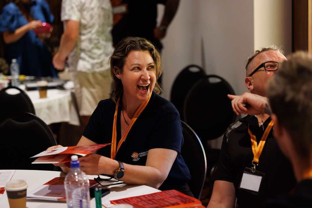 A smiling person sits at a table with others, holding open a red document while a colleague points toward something nearby.