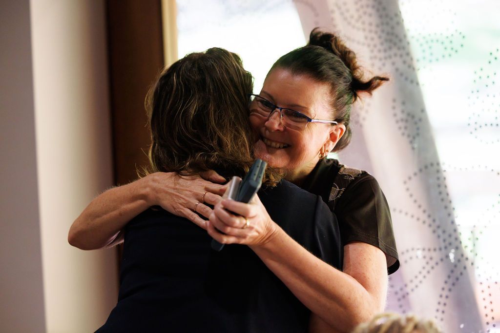 A person wearing glasses smiles warmly while hugging another individual in a sunlit indoor setting.