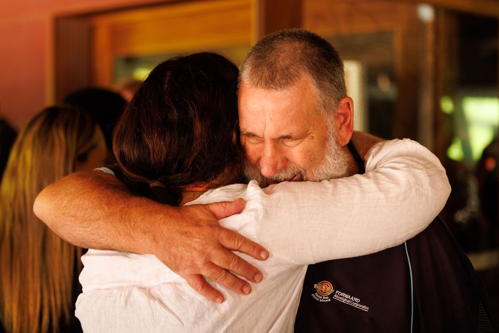 A man with a white beard and a black shirt embraces another person in a white top, their faces pressed together.
