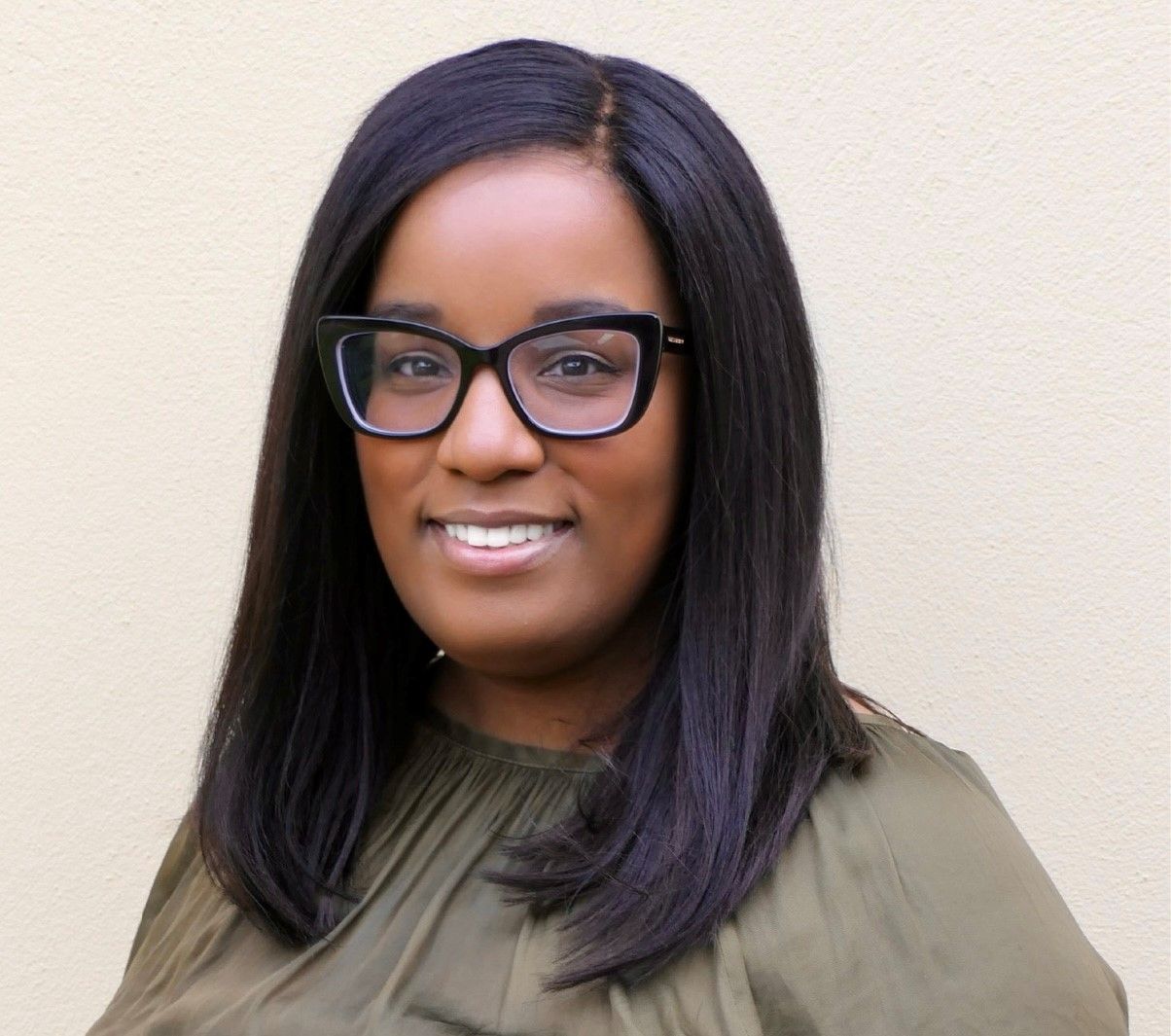A person with dark hair and glasses wearing an olive green top, smiling against a neutral wall.