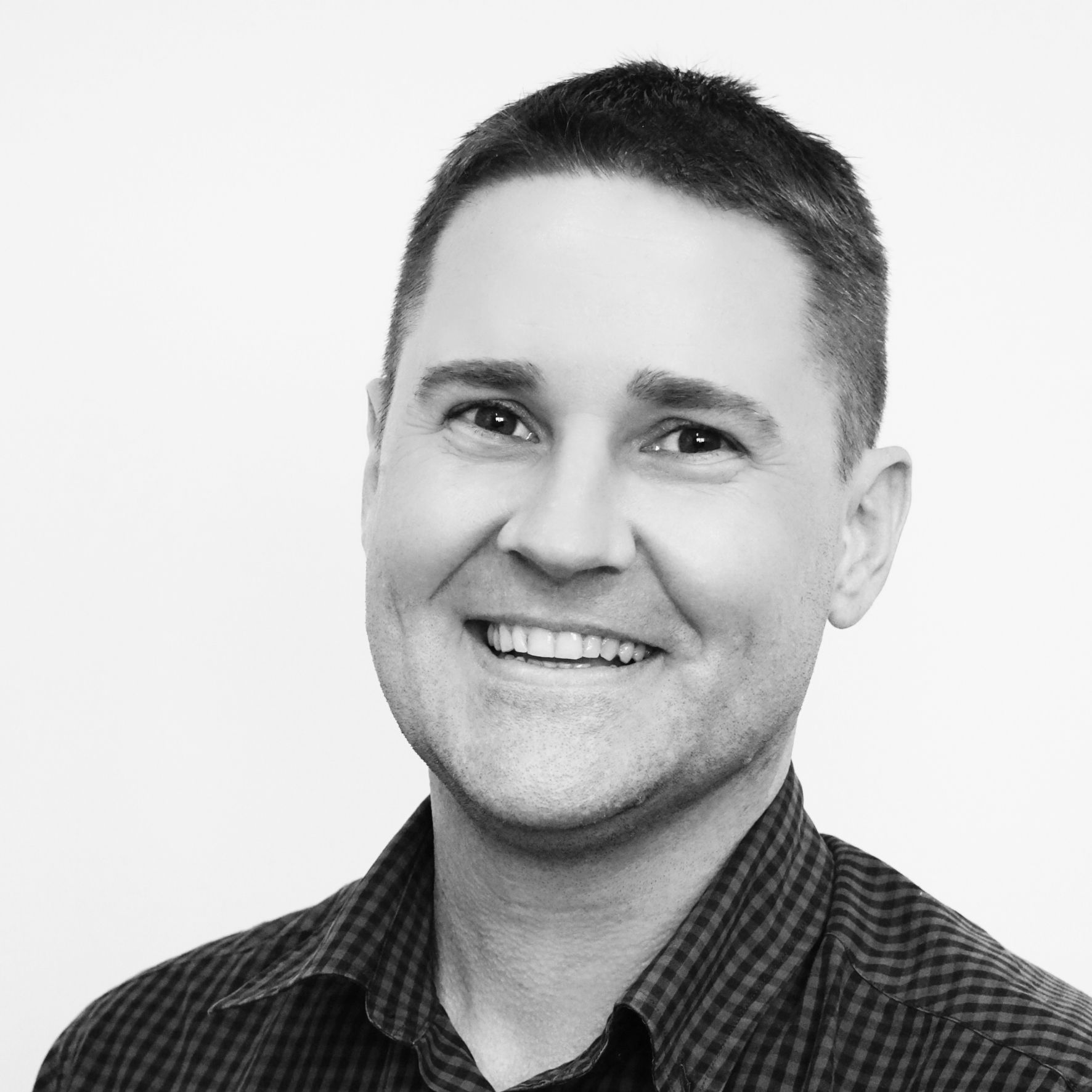 A smiling person in a dark-colored patterned button-down shirt, captured in a black and white headshot against a light wall.