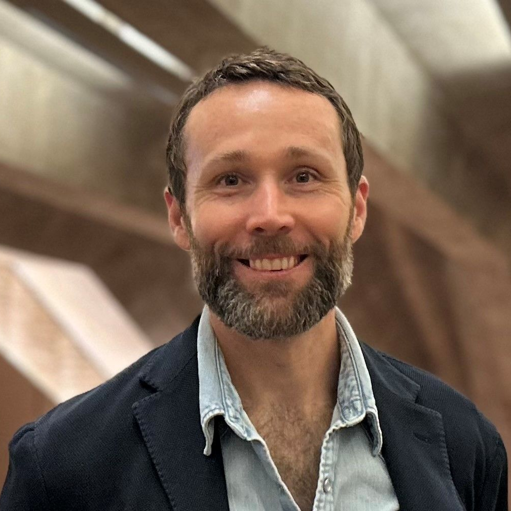 A person with a beard, wearing a light blue collared shirt under a dark blazer, smiles against a blurred backdrop.