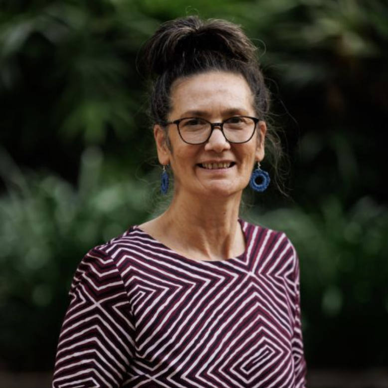 A person with their hair in a high bun wears glasses and blue circular earrings, posing against a blurred green background.
