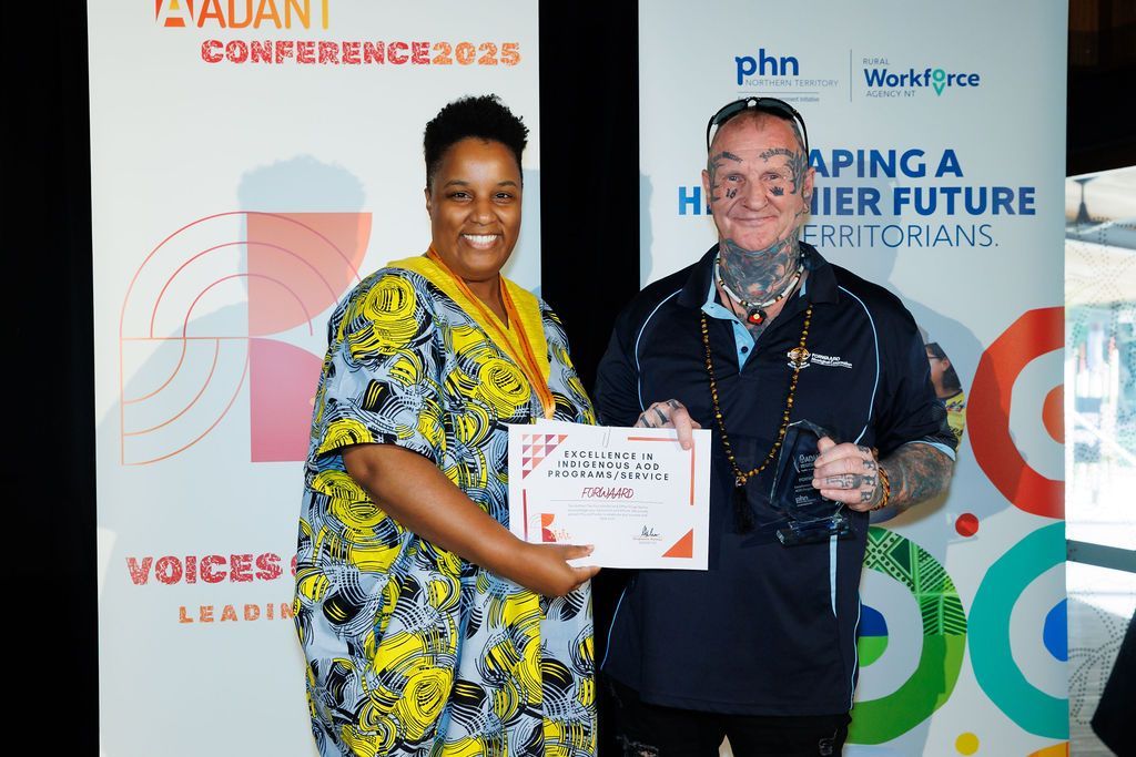 Two people stand smiling in front of a conference banner, holding an award certificate at an awards ceremony.