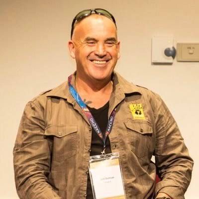 A smiling person with sunglasses perched on their head wears a tan button-up shirt over a black top and a conference badge.