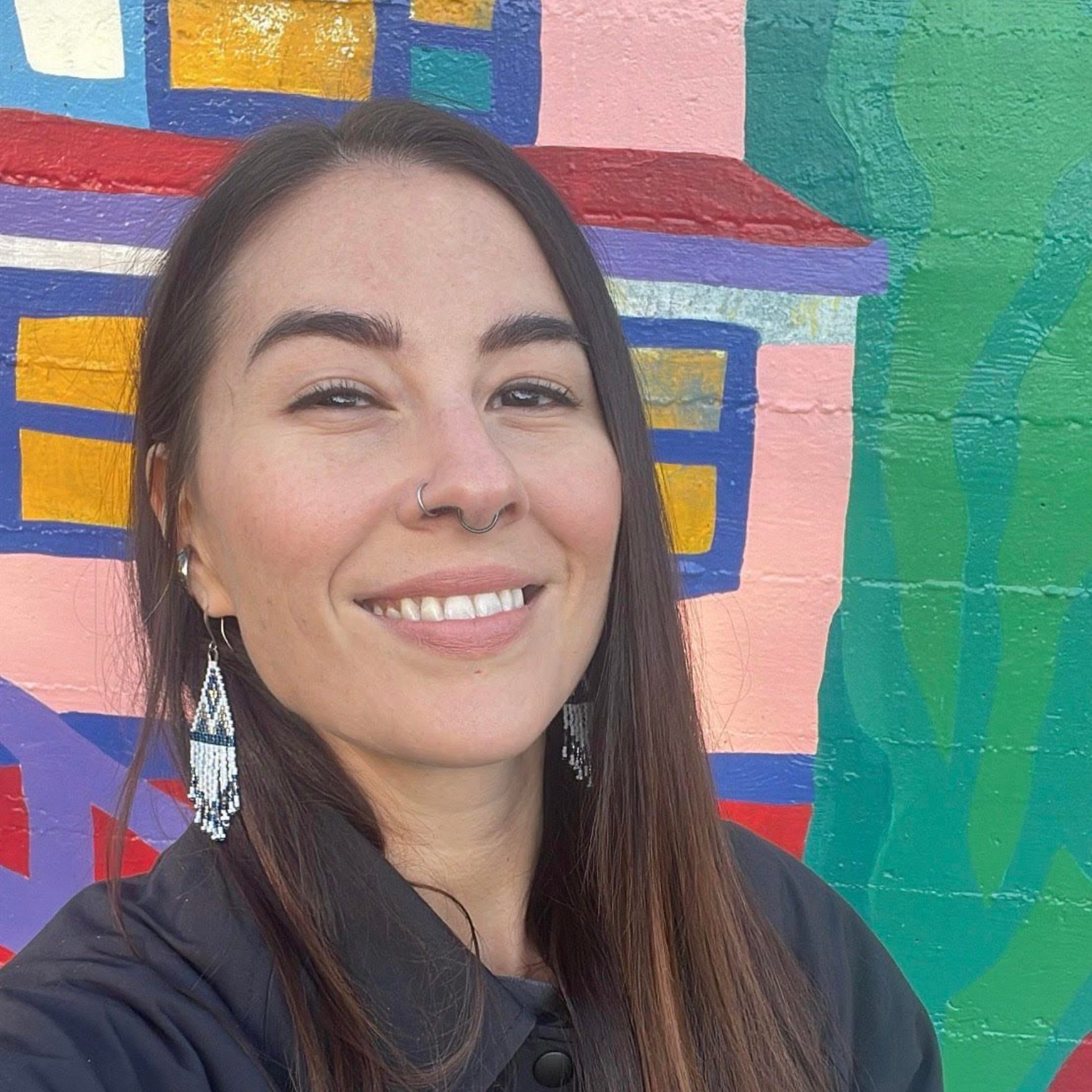 A smiling person with long dark hair, wearing dangling earrings and a nose ring, stands in front of a colorful wall mural.
