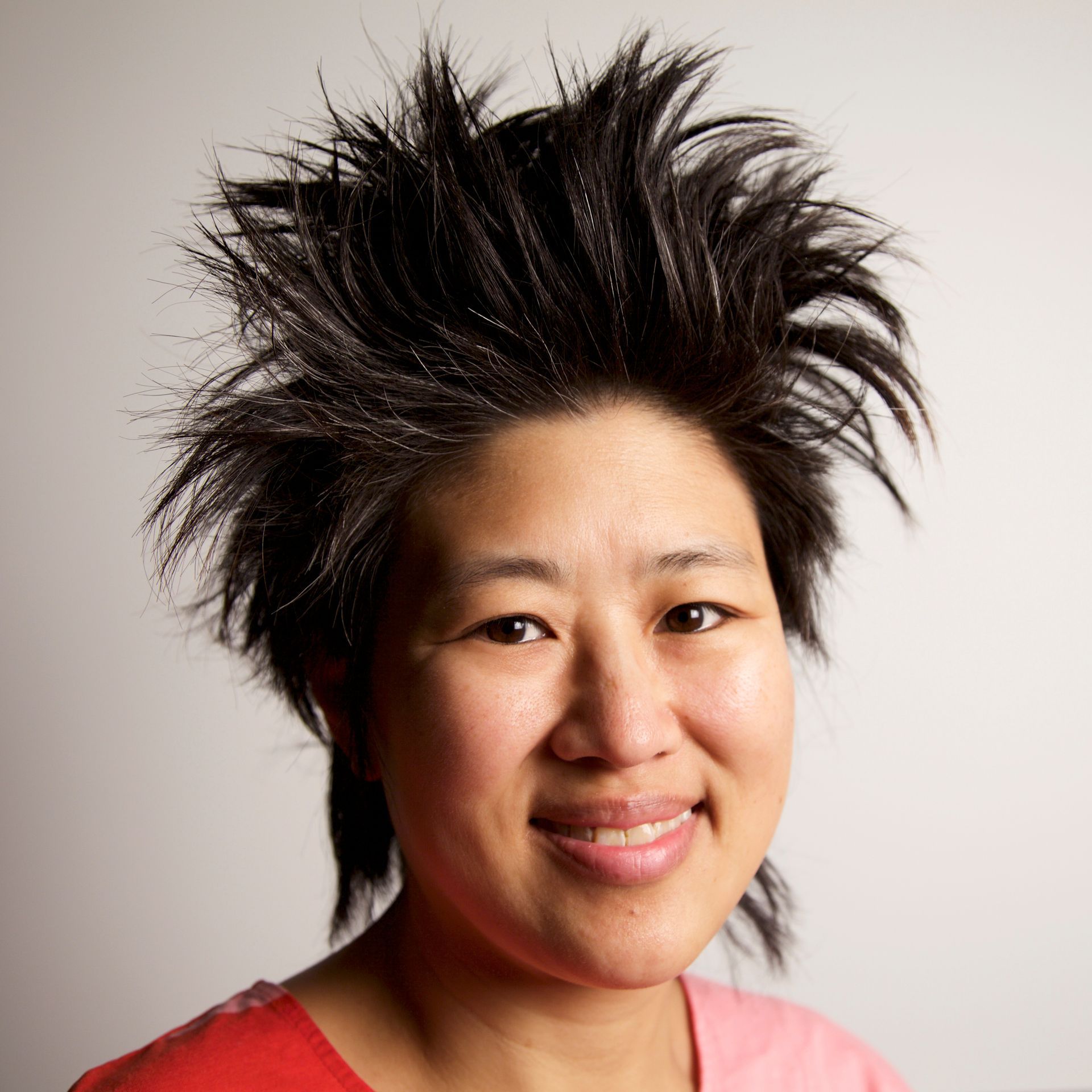A smiling person with dark, short hair styled in a high, textured, spiky fashion against a plain light gray background.