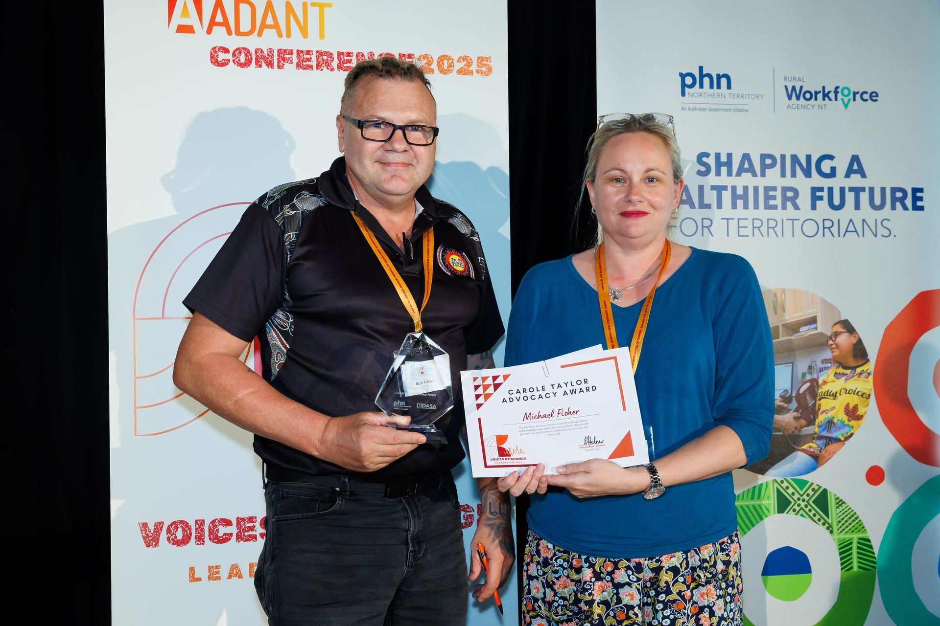 A person with glasses holding an award and a person holding a certificate stand before two conference banners.