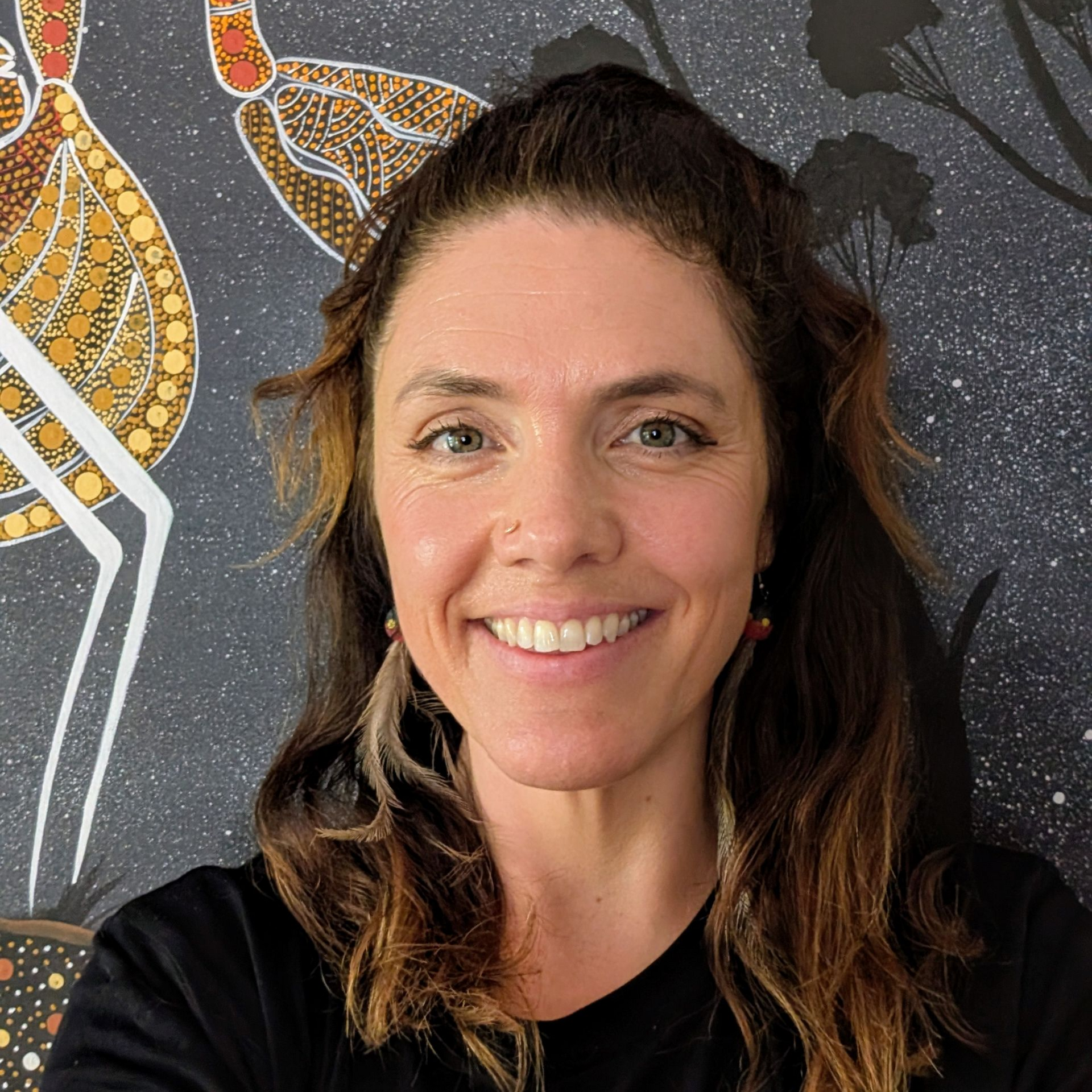 A smiling person with wavy brown hair wearing feather earrings, centered against a dark patterned art backdrop.