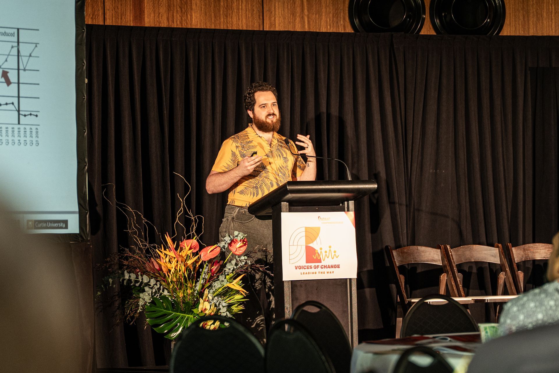 A person with a beard in a patterned yellow shirt speaks at a podium in front of a dark curtain.