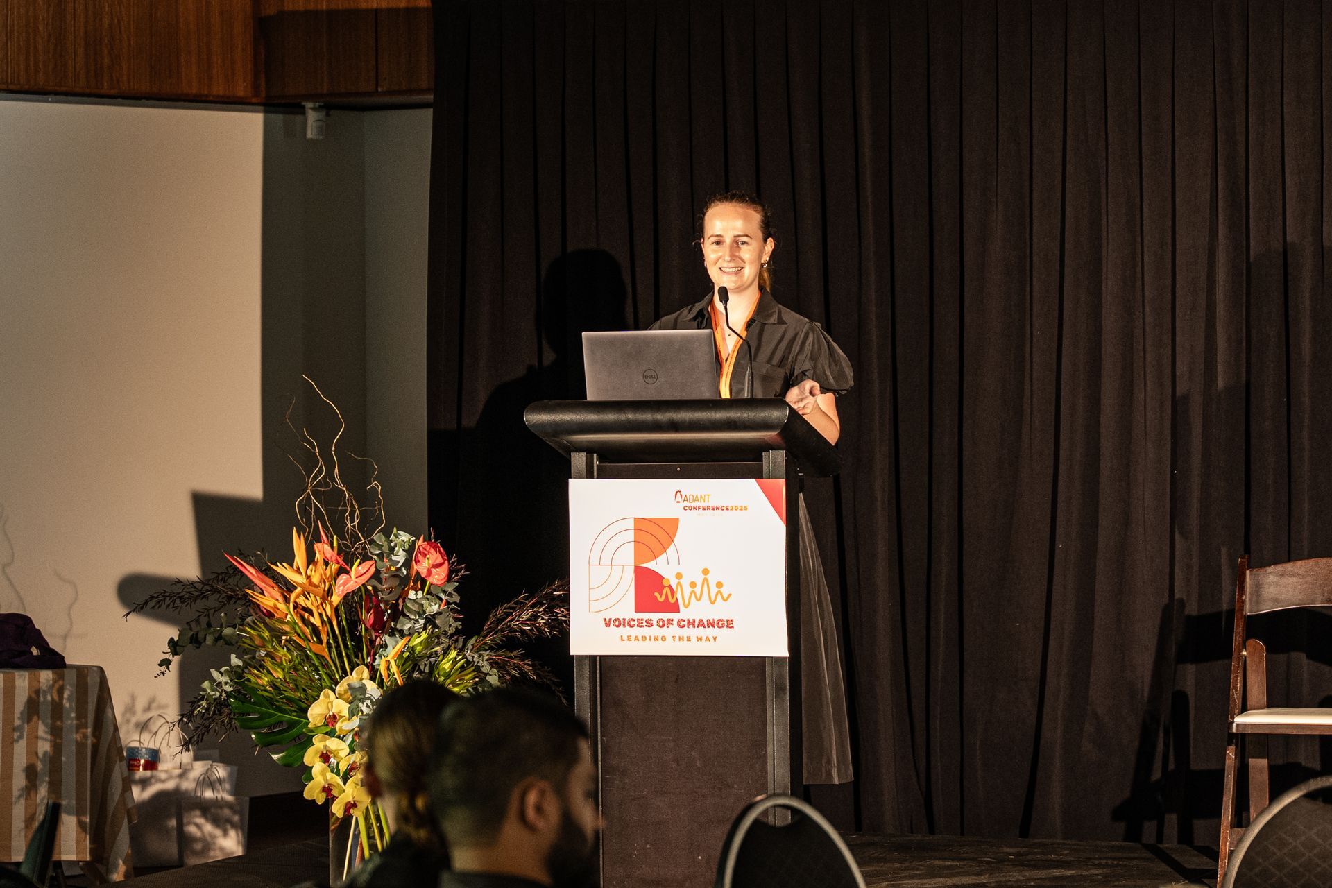 A presenter stands at a podium with a laptop, speaking at a conference event in front of a dark curtain backdrop.