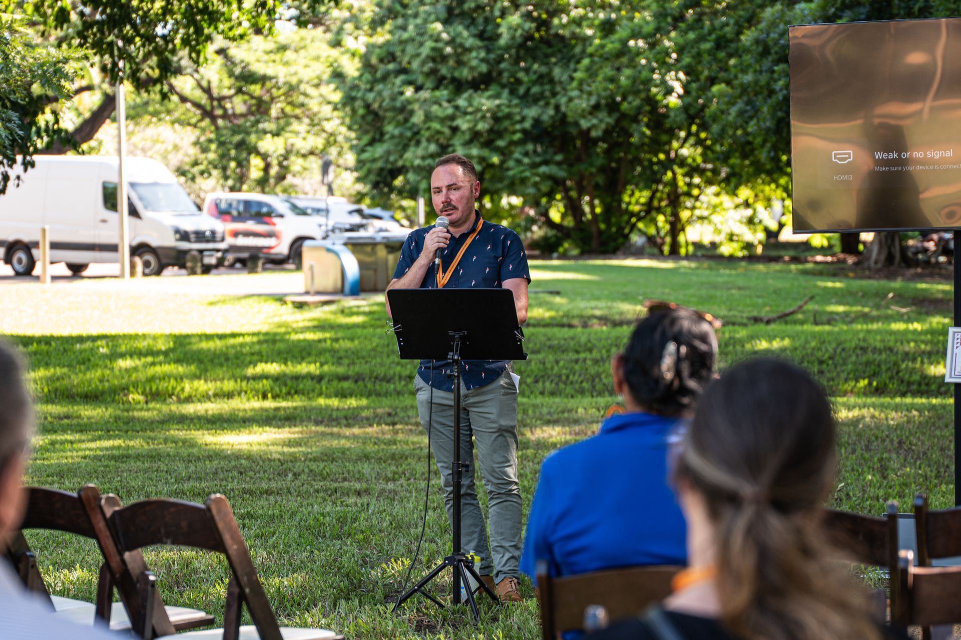 A person speaks into a microphone at a podium outdoors, facing a seated audience in a green, tree-lined park setting.