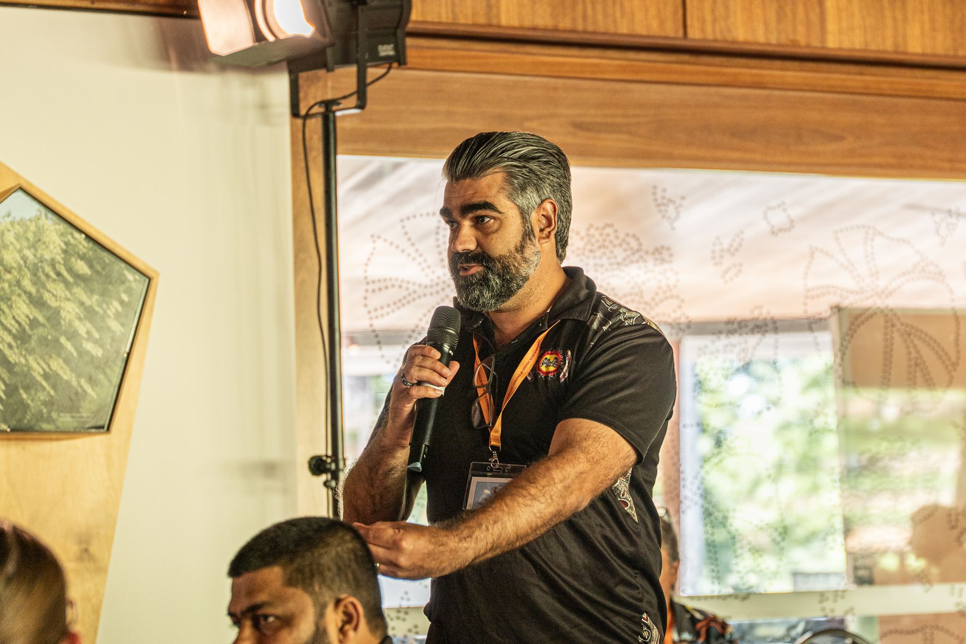 A man with a beard holds a microphone while speaking at an indoor event, standing next to a person in the foreground.