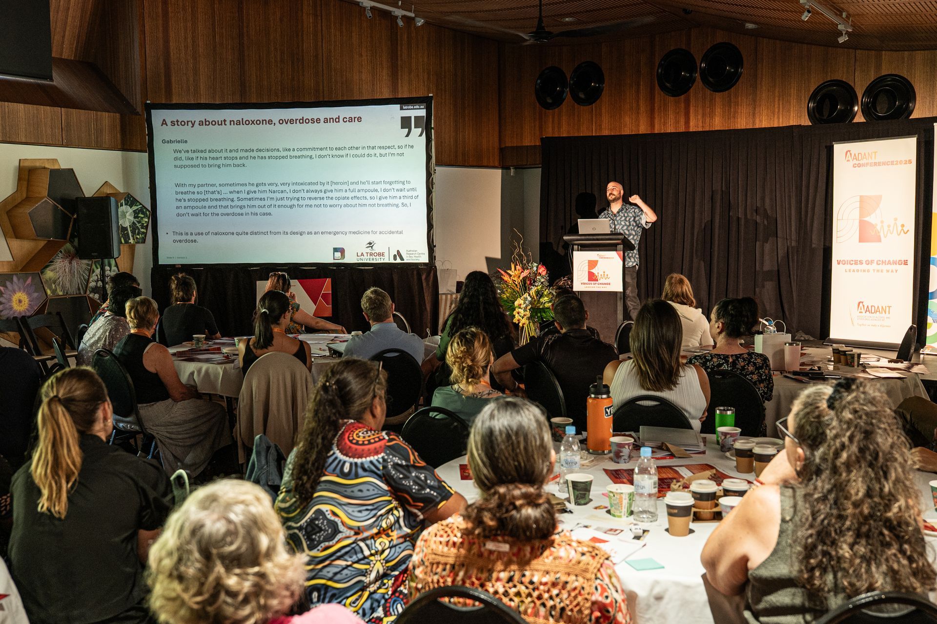 A speaker presents to an audience seated at tables in a wood-paneled conference room with a large projection screen.