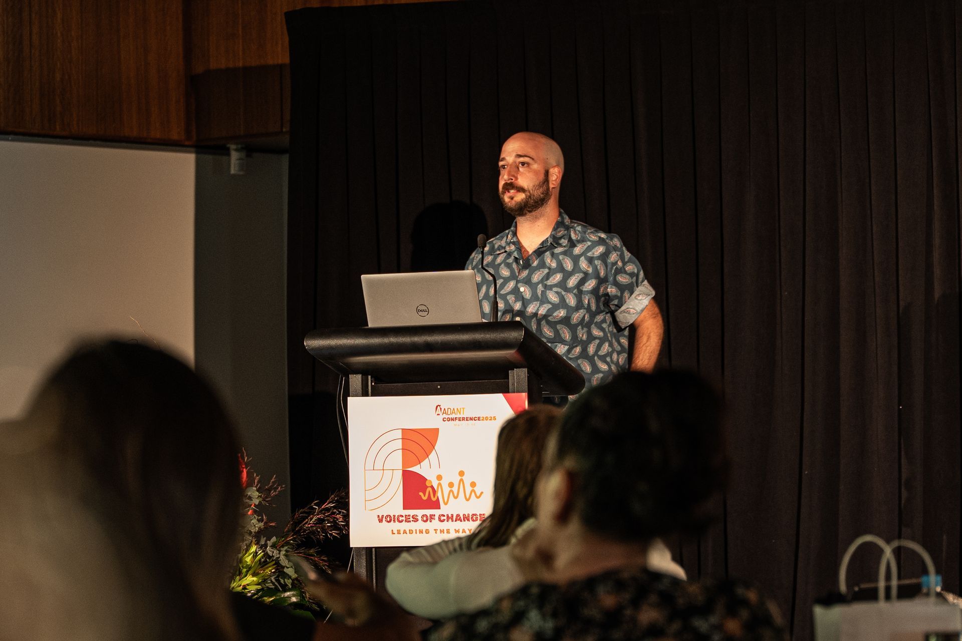 A presenter stands at a lectern with a laptop in front of an audience in a dimly lit room with black curtains.