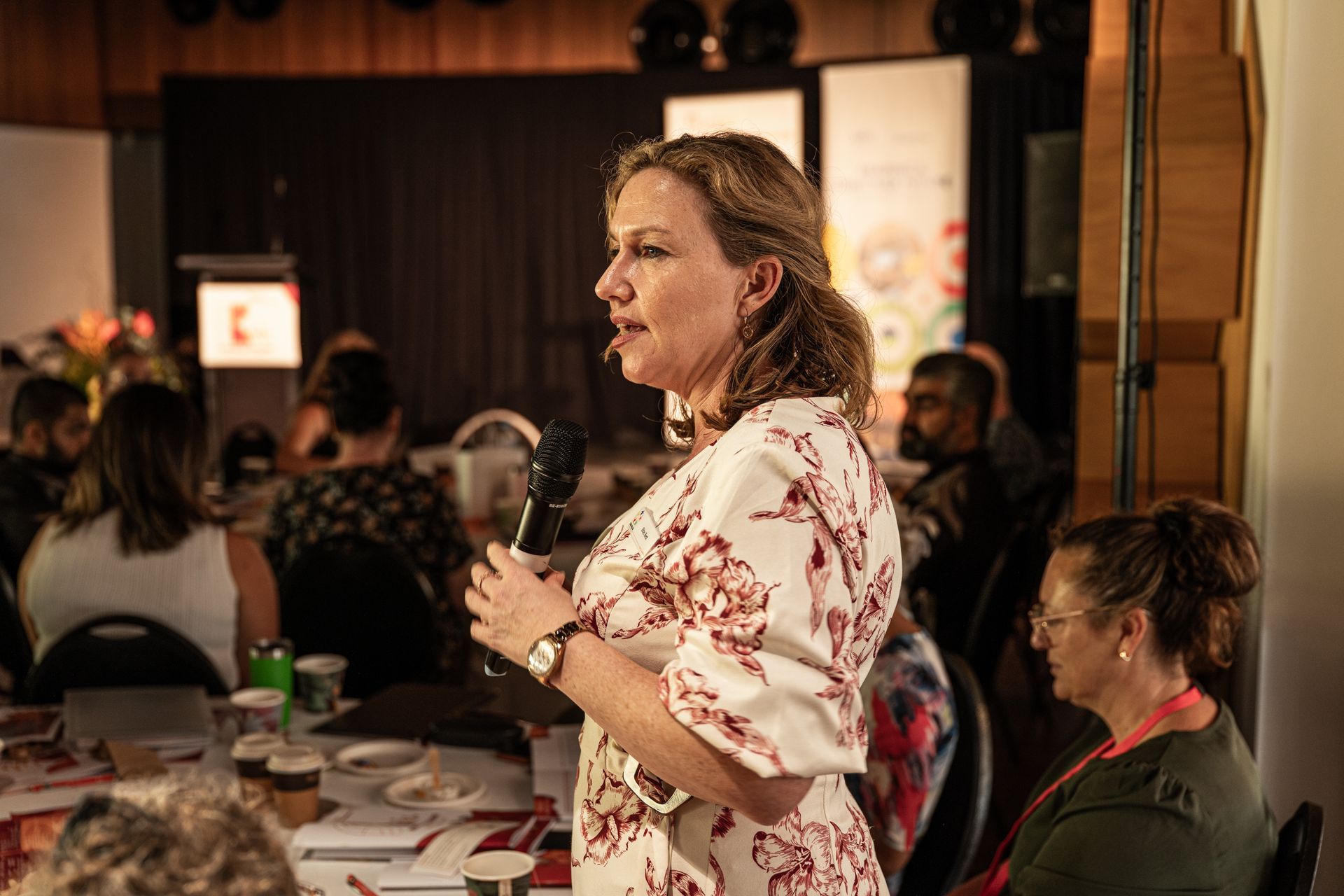 A speaker in a white floral top holding a microphone addresses an audience in a dim indoor conference setting.