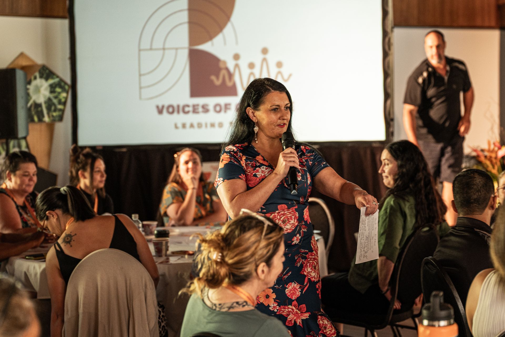 A speaker in a floral dress addresses a group at a conference, holding a small paper in front of a branded screen.