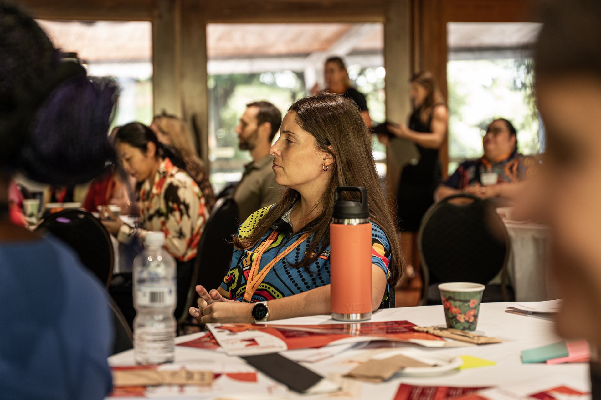 A group of people listens attentively at a brightly lit indoor workshop with tables, printed materials, and a water bottle.