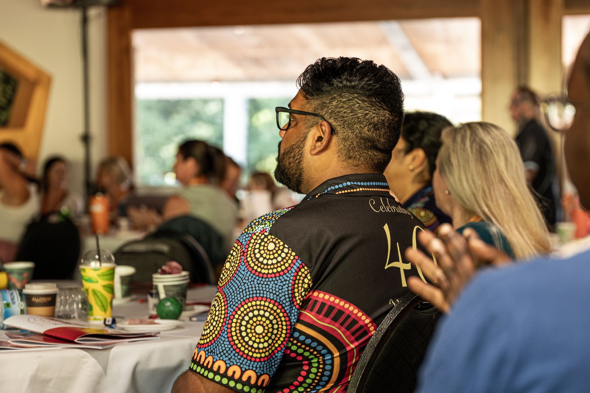 A side view of a person in a patterned shirt clapping at a conference, with other attendees seated at tables nearby.
