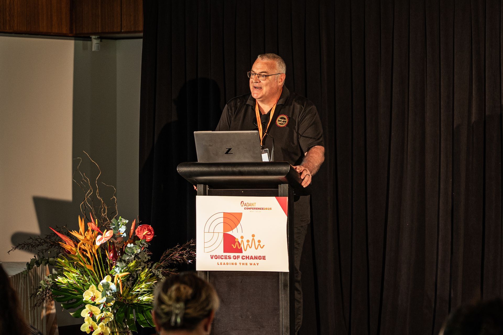 A person stands at a podium with a laptop, speaking at an event in front of black curtains and a large floral arrangement.