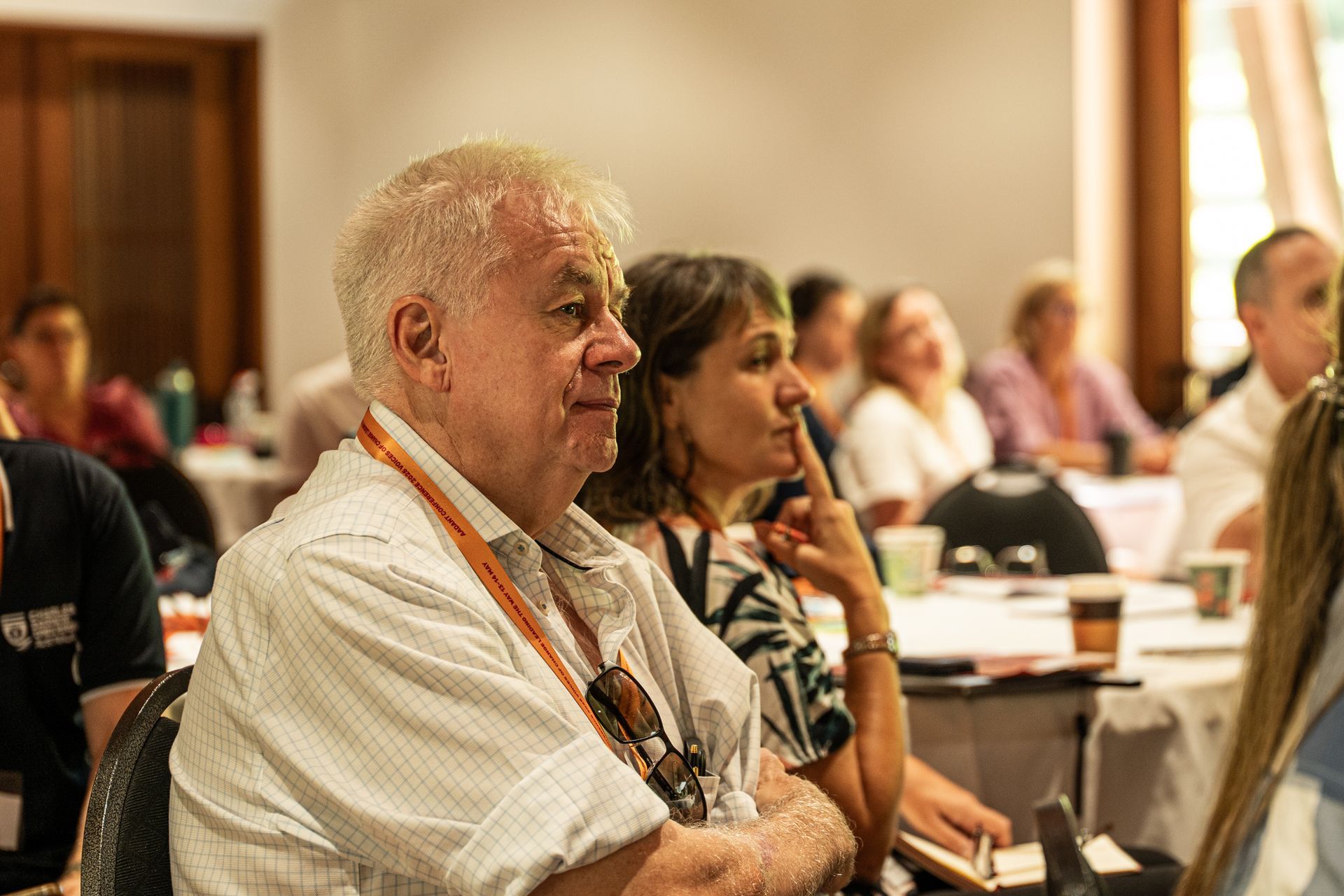 A group of people sit at tables in a room, listening to a presentation during a conference or meeting.