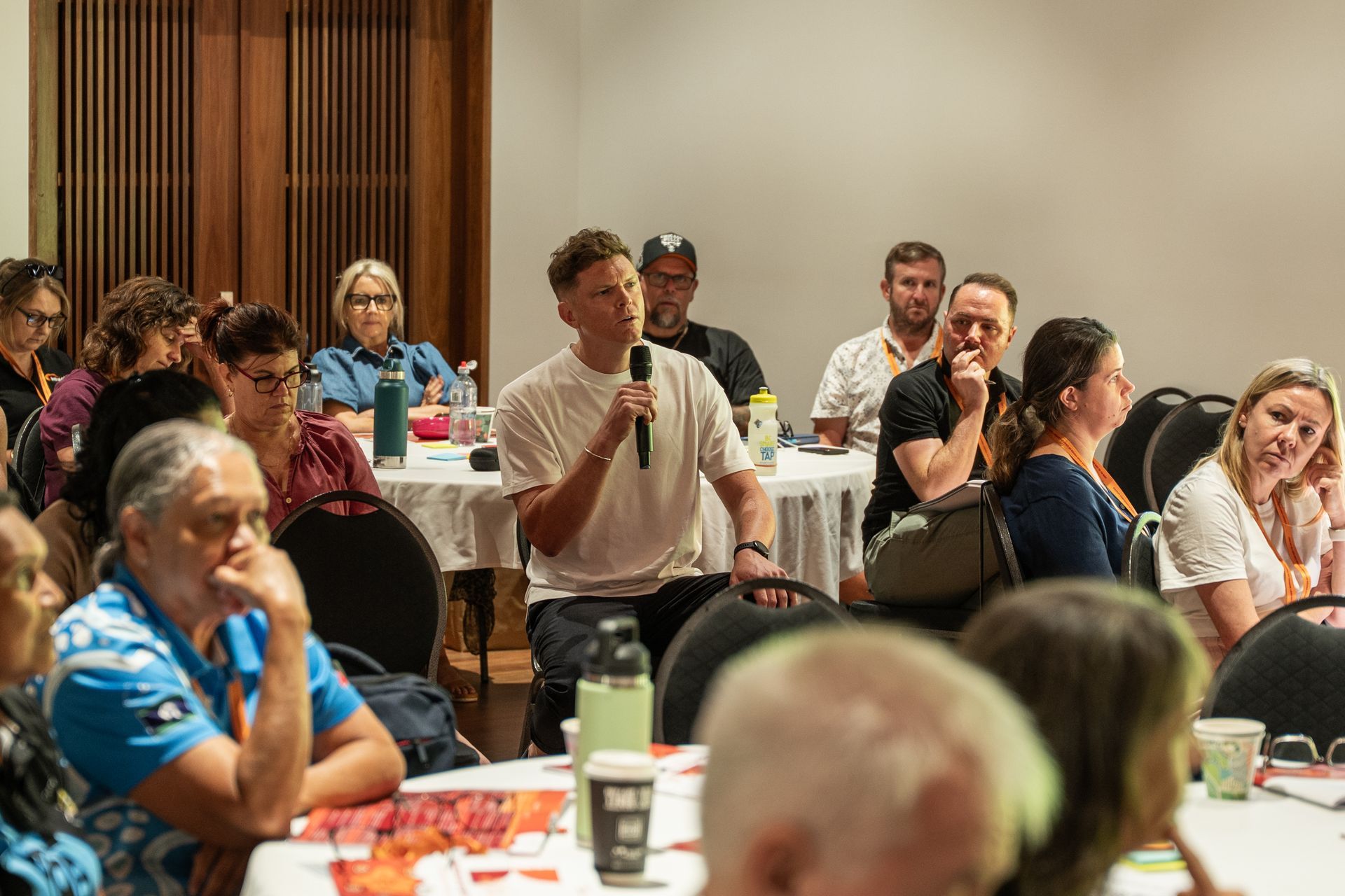 A person speaks into a microphone during a meeting as others listen at tables in a brightly lit room.