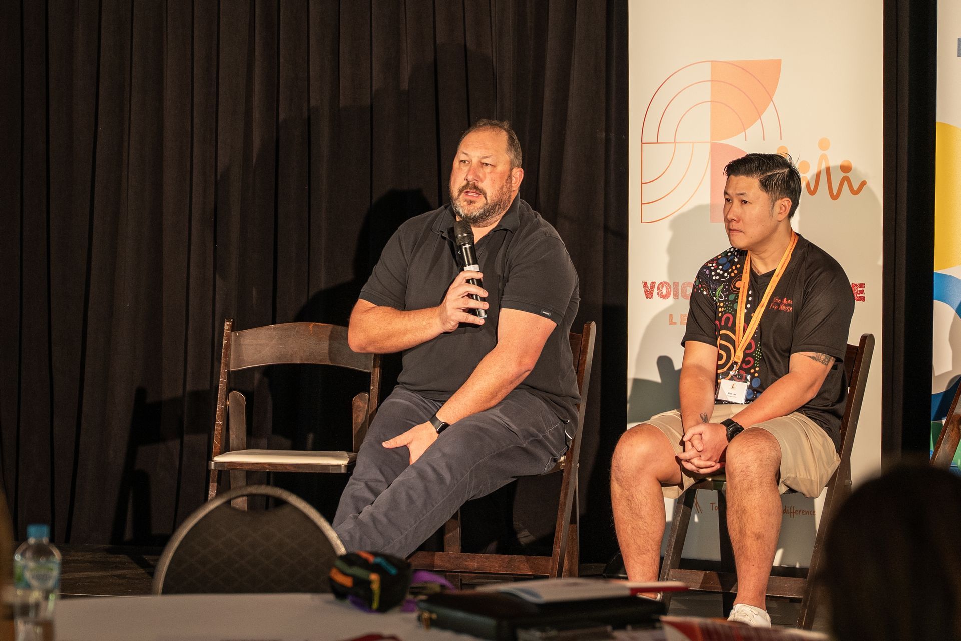 Two presenters sit on stage in chairs before a dark curtain, with one holding a microphone to speak.