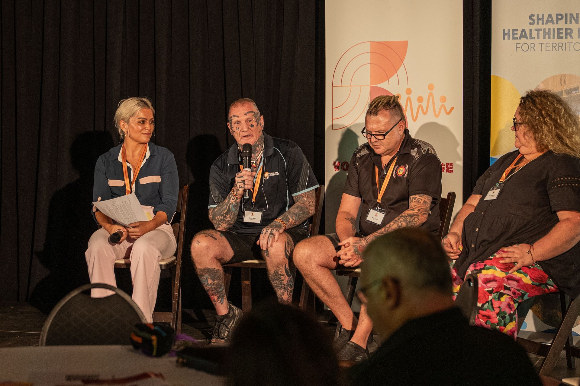 Four people sit in a row on chairs on a stage during a presentation, with one person speaking into a microphone.