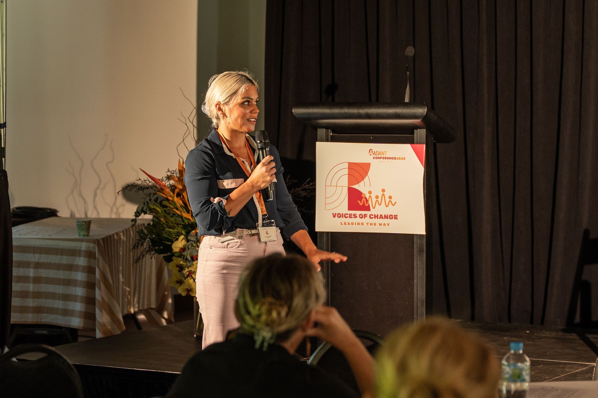 A person stands at a podium with a microphone, presenting to an audience in a room with a curtained background.