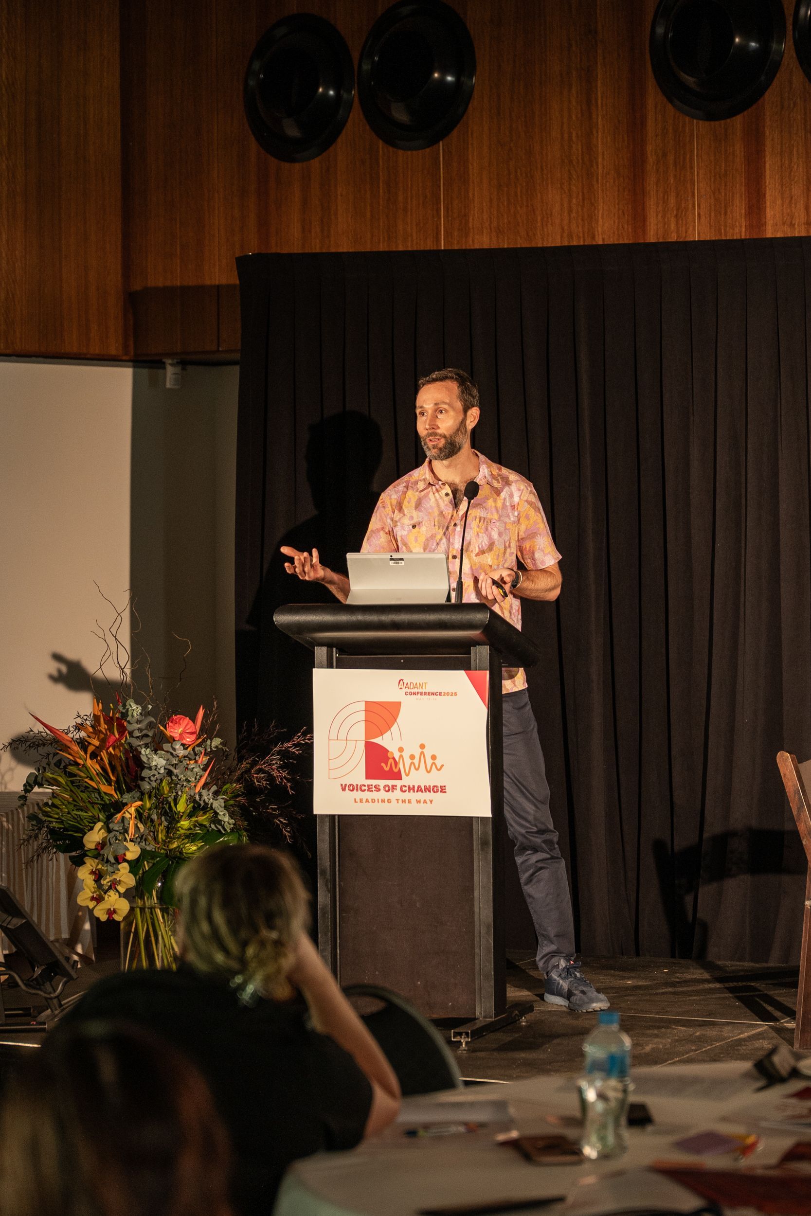 A person in a patterned shirt stands behind a podium speaking to an audience in a dimly lit auditorium.