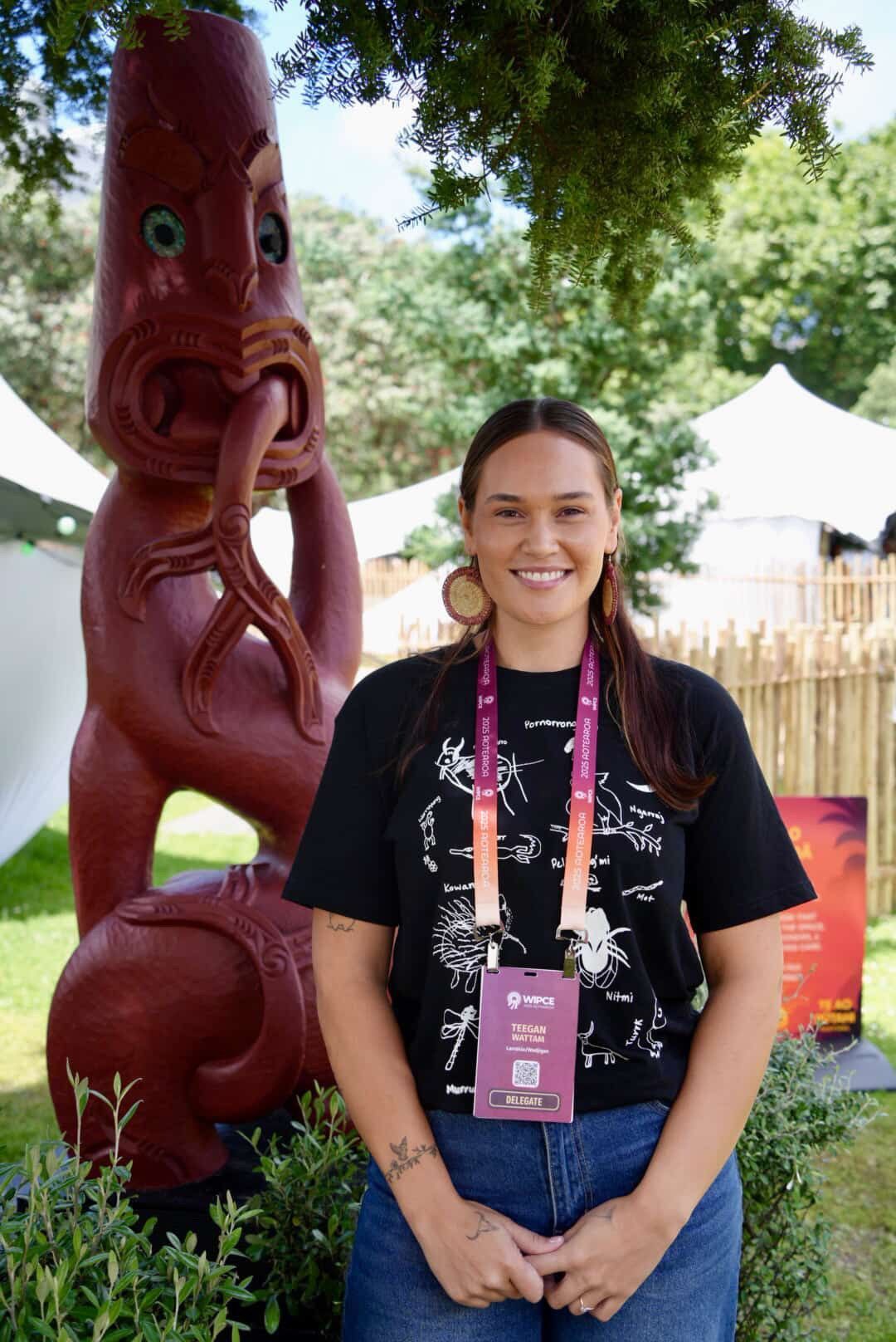 A person smiling while standing in front of a red carved wooden figure in an outdoor park setting.