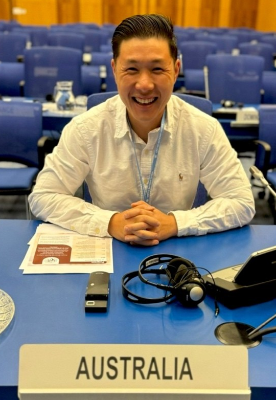 A smiling individual sits at a blue conference table behind an Australia placard, with a headset and papers nearby.
