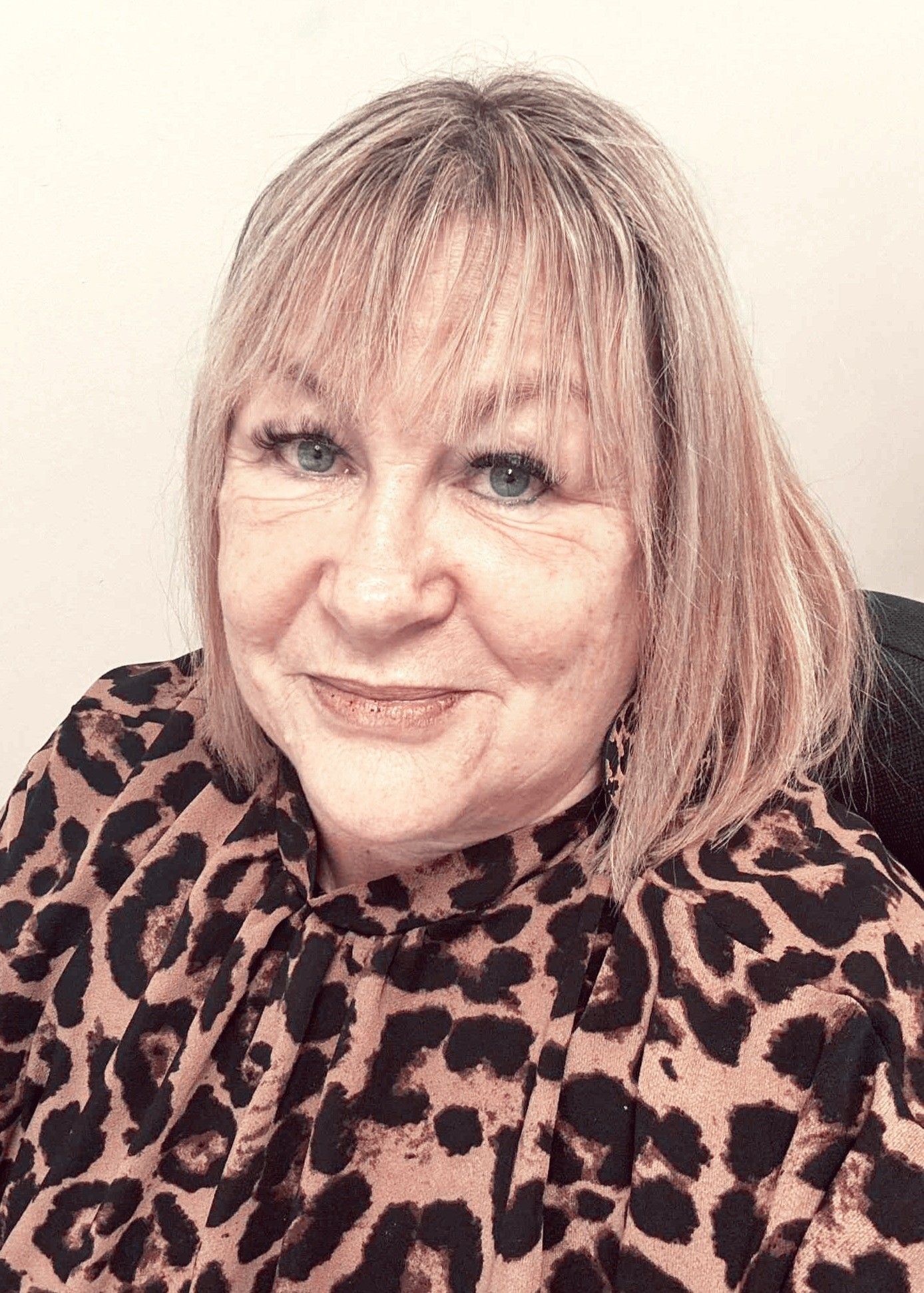 A person with blonde hair and bangs, wearing a brown and black leopard-print top, smiles slightly at the camera.