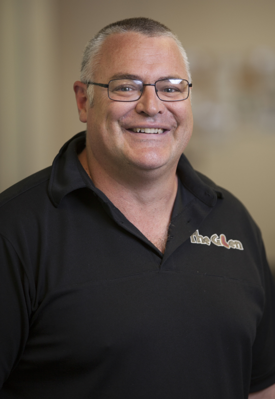 A person with short hair and glasses wearing a black polo shirt, smiling at the camera in a soft-focus indoor setting.