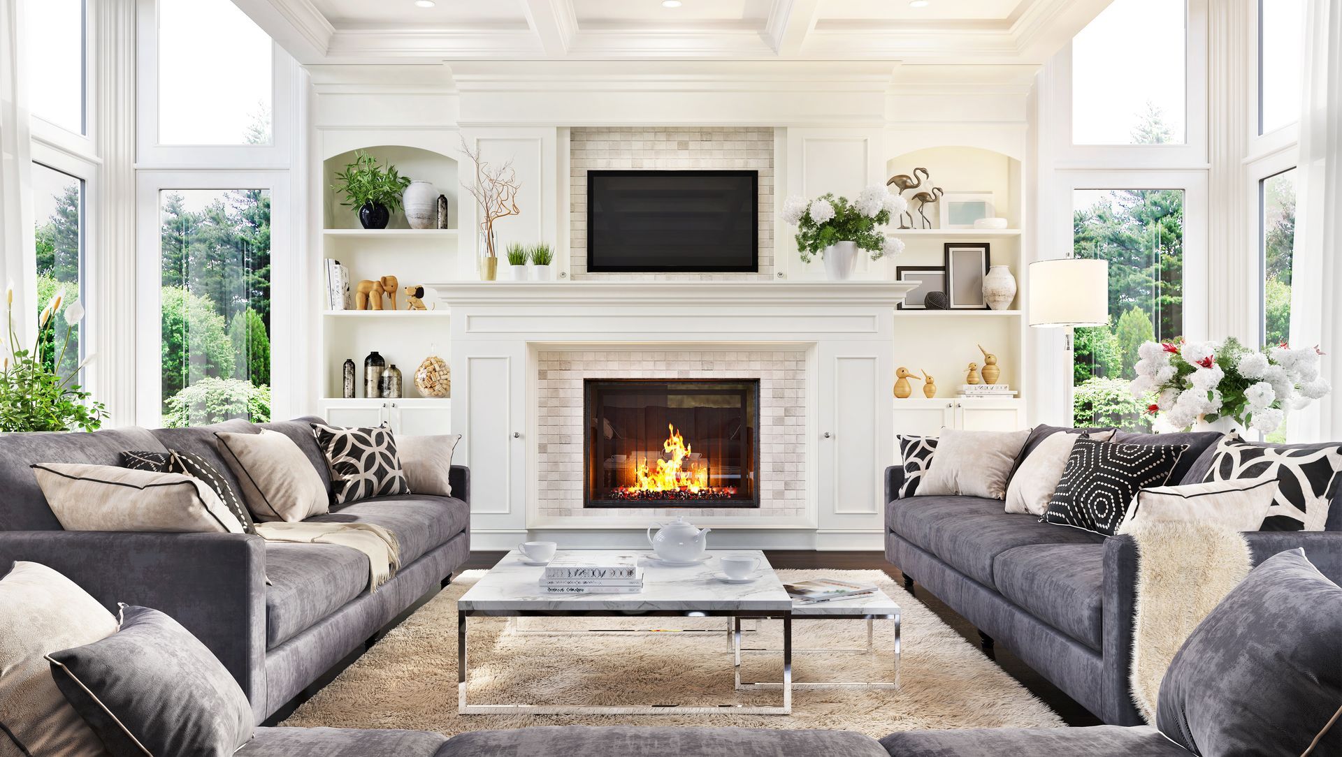 Spacious living room with gray couches, fireplace, and large windows overlooking greenery.