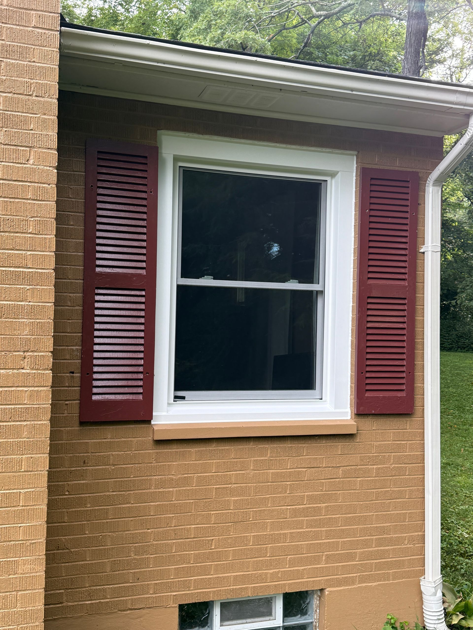 Window with red shutters on a brick wall, under white trim and a green roof.