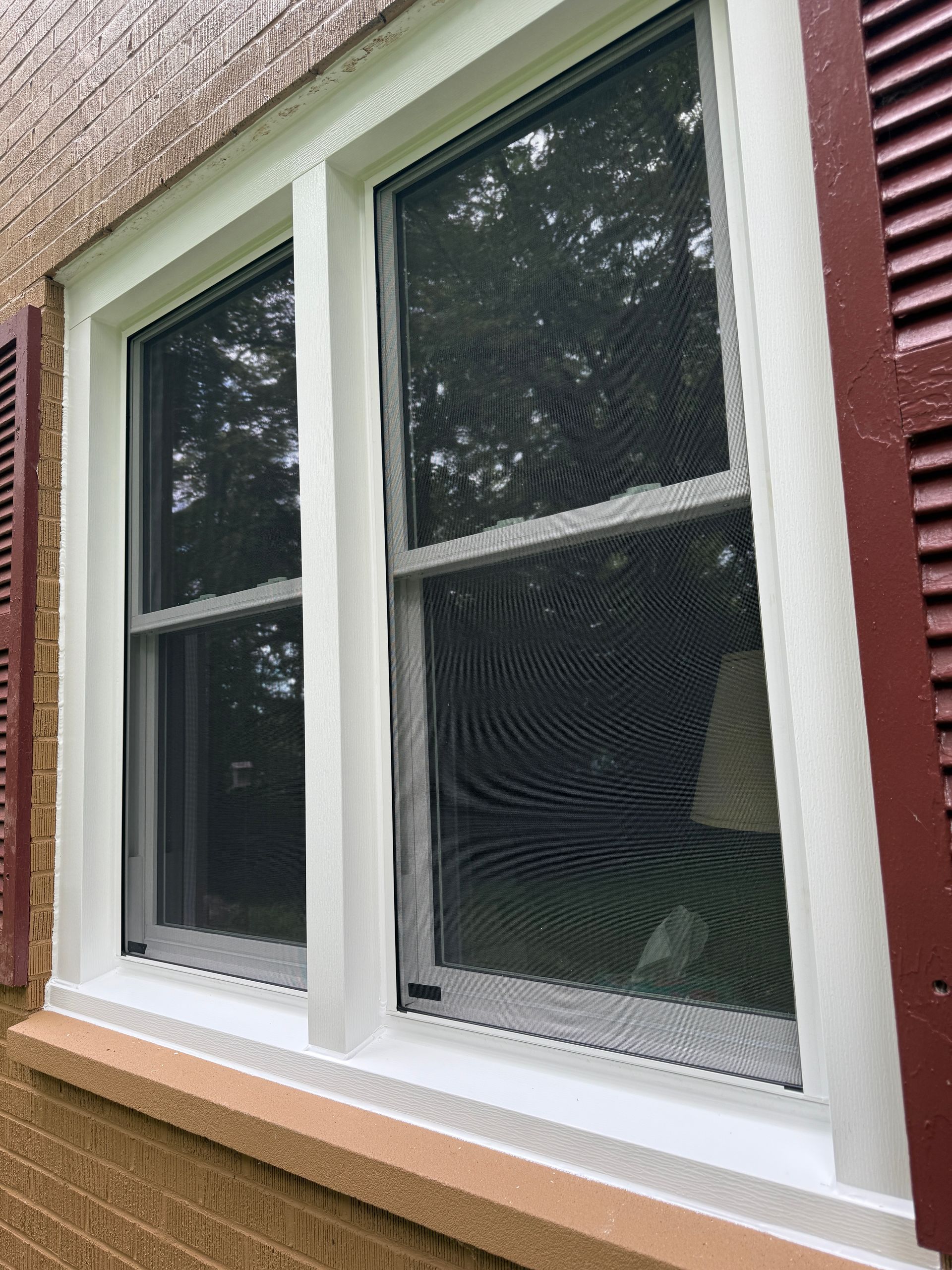 Two white-framed, double-hung windows with black screens and red shutters against a brick exterior.
