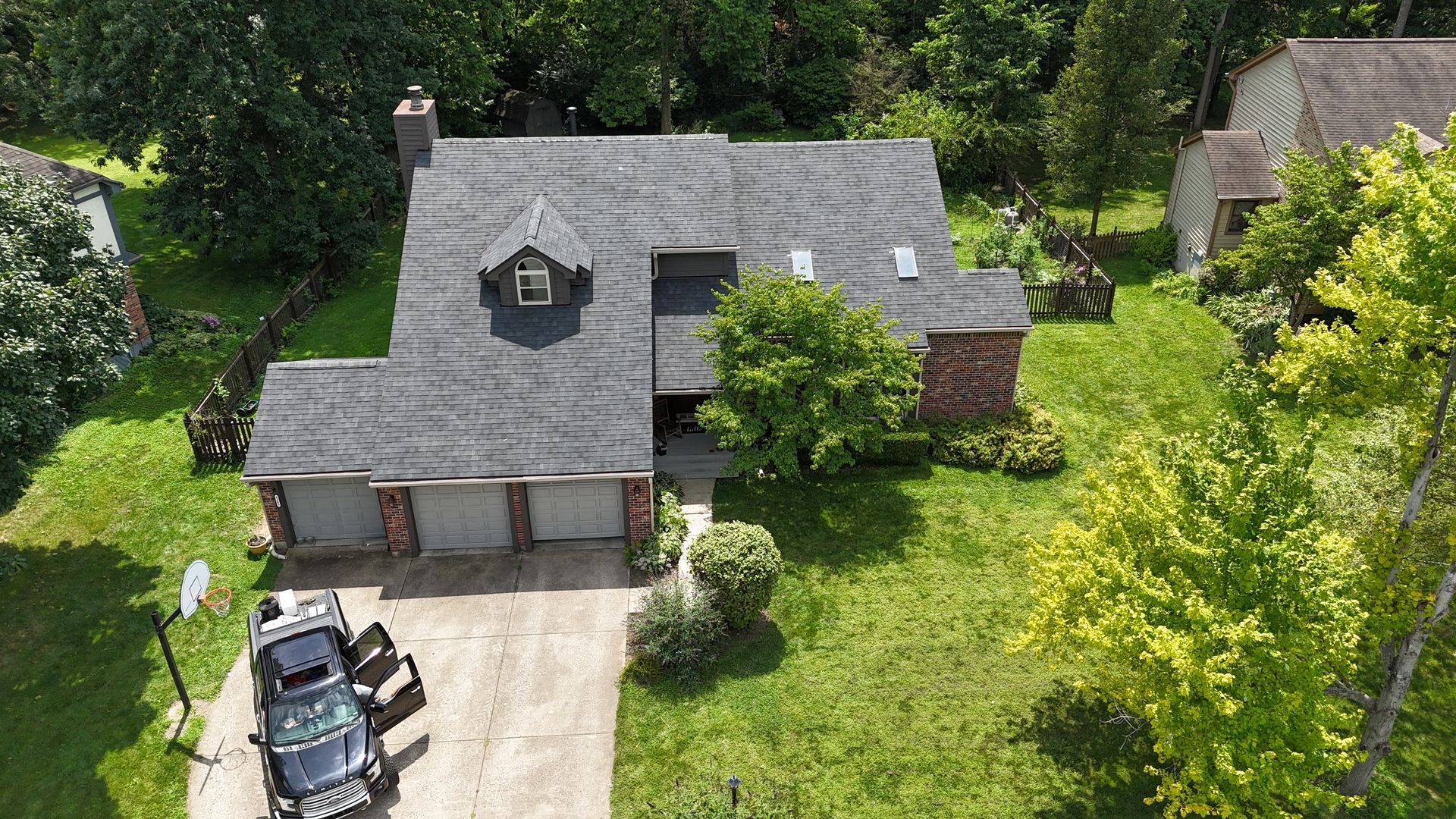 Aerial view of a gray-roofed house with three garage doors and a car parked in the driveway, surrounded by green trees and grass.