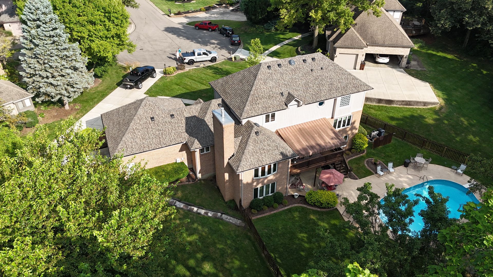 Aerial view of a large house with a pool and cars in the driveway, surrounded by green trees and grass.
