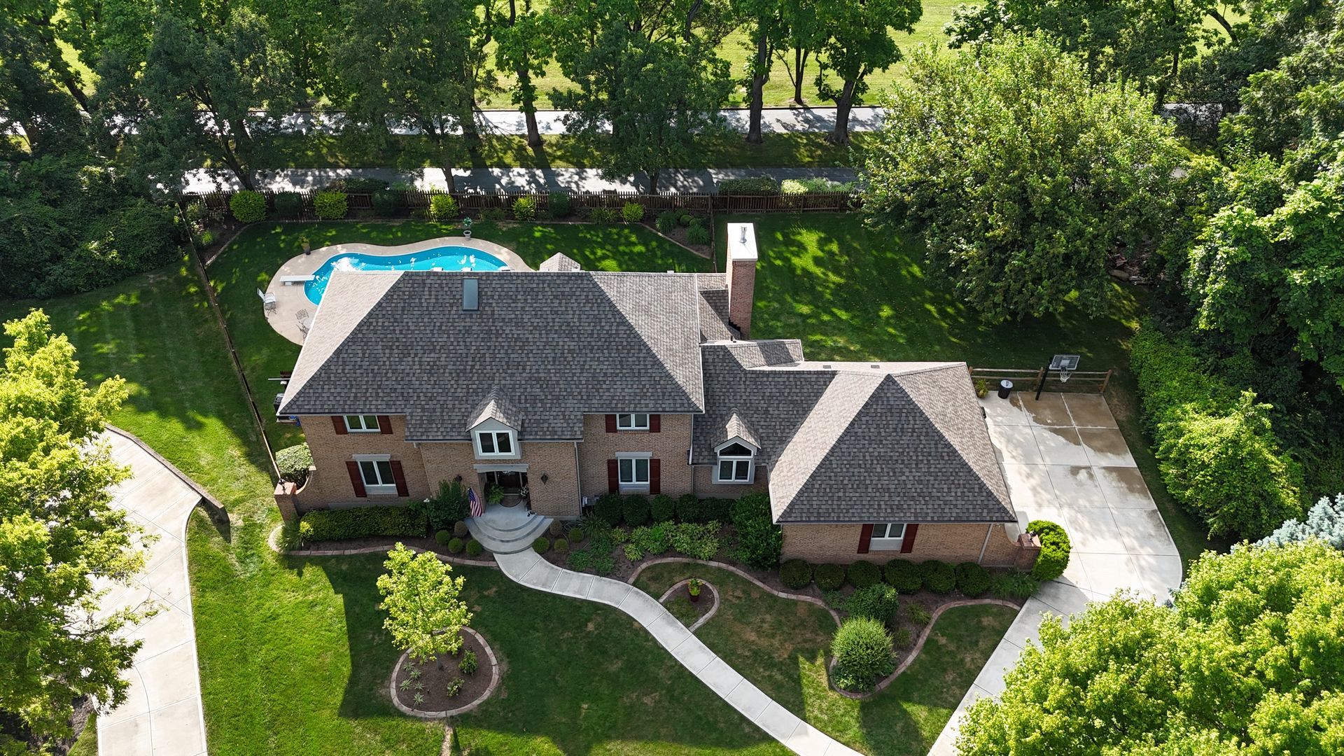 Aerial view of a large brick house with a pool and long driveway, surrounded by trees and green lawn.