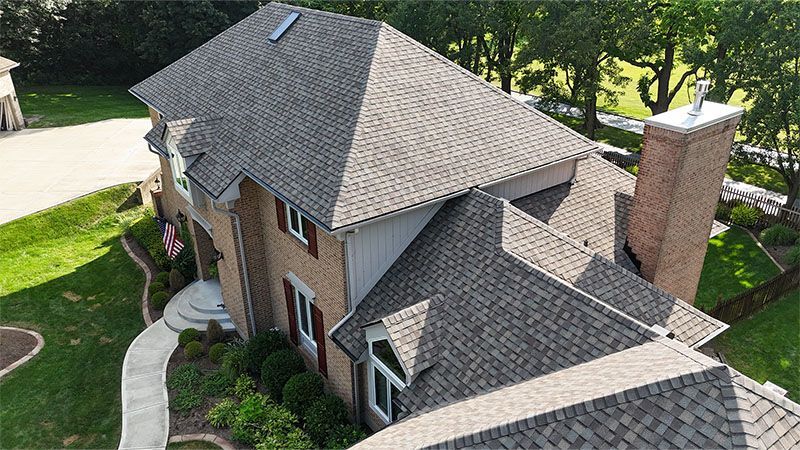 Aerial view of a two-story brick house with a complex shingled roof, chimney, and green lawn.