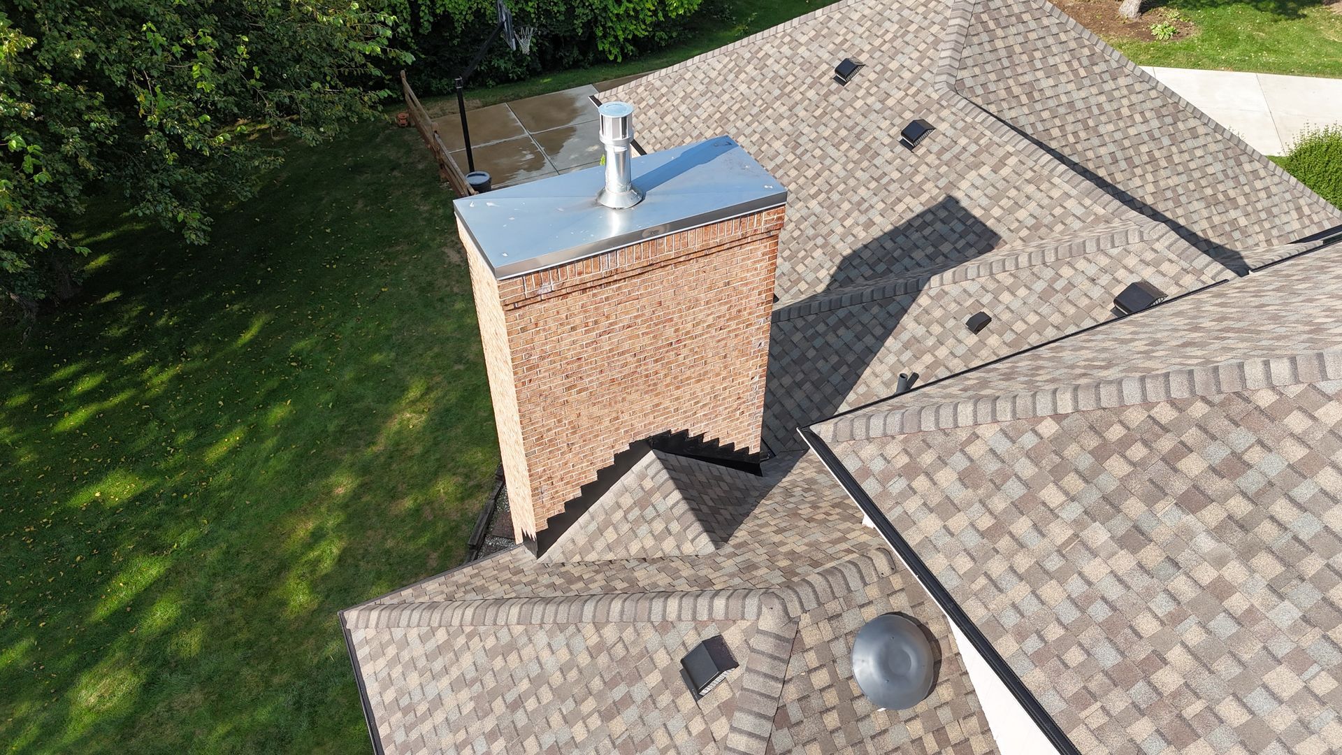 Overhead view of a house roof with a brick chimney and vent, covered in brown asphalt shingles.