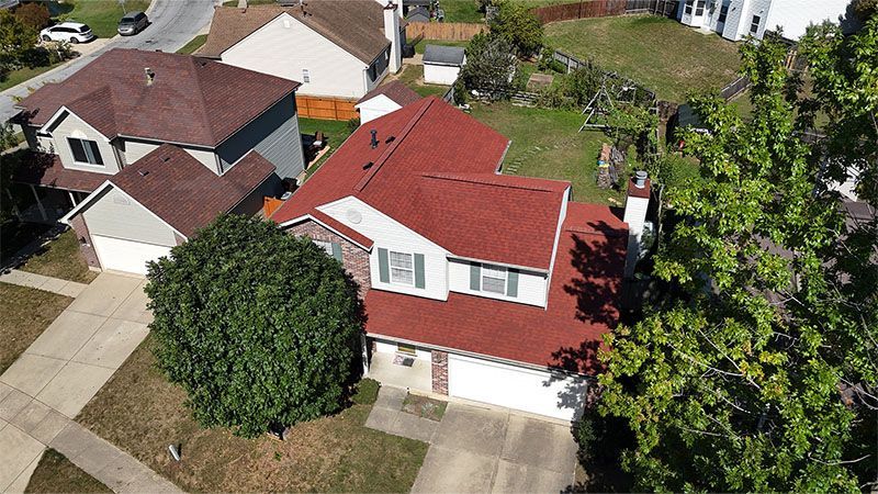 Aerial view of a suburban house with a red roof, white trim, and a large green tree in the front yard.
