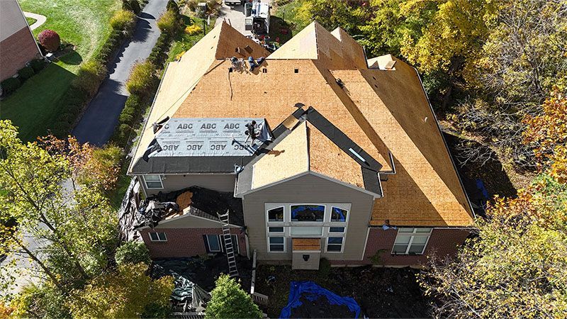 Aerial view of a house undergoing roof replacement; workers on roof; exposed wood, shingles, and building materials visible.